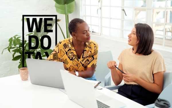 Two individuals seated at a table with laptops, engaged in a discussion. A plant and a multi-paned window are visible in the background, creating a collaborative and professional work setting in Australia.